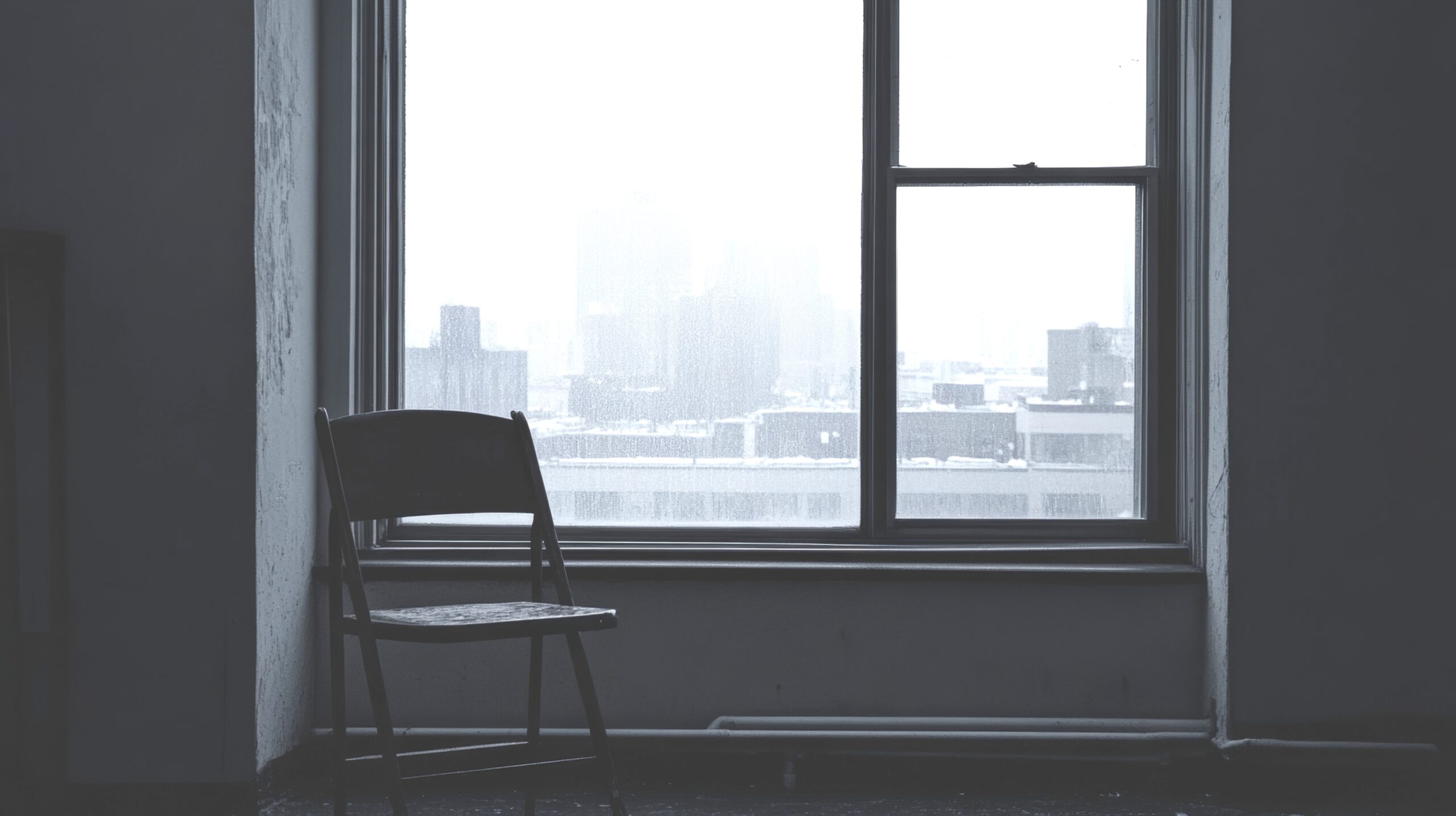 Empty room with a solitary chair facing a foggy city view through a window. The gray scene evokes a sense of loneliness and contemplation.