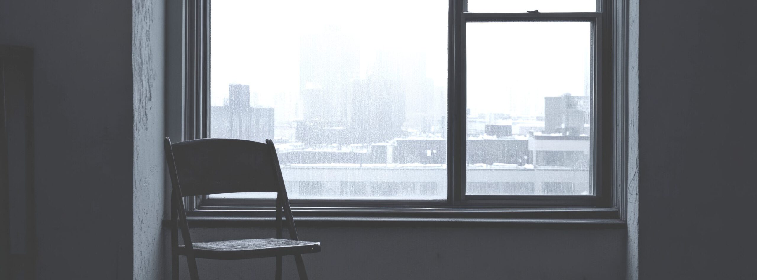 Empty room with a solitary chair facing a foggy city view through a window. The gray scene evokes a sense of loneliness and contemplation.
